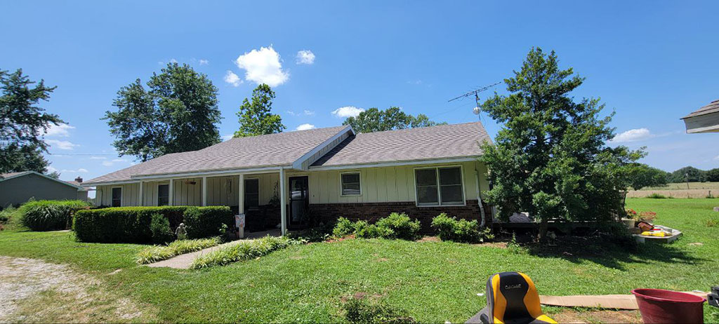 White ranch house with a new light gray shingle roof, trees and lawn, Dedicated Builders