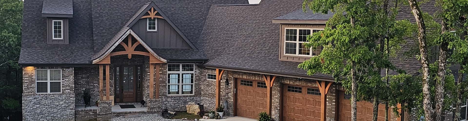 Craftsman-style brick and stacked-stone house with a dark architectural shingle roof broken by two dormers, front gables and valleys, and a timber-bracketed entry porch, Dedicated Builders