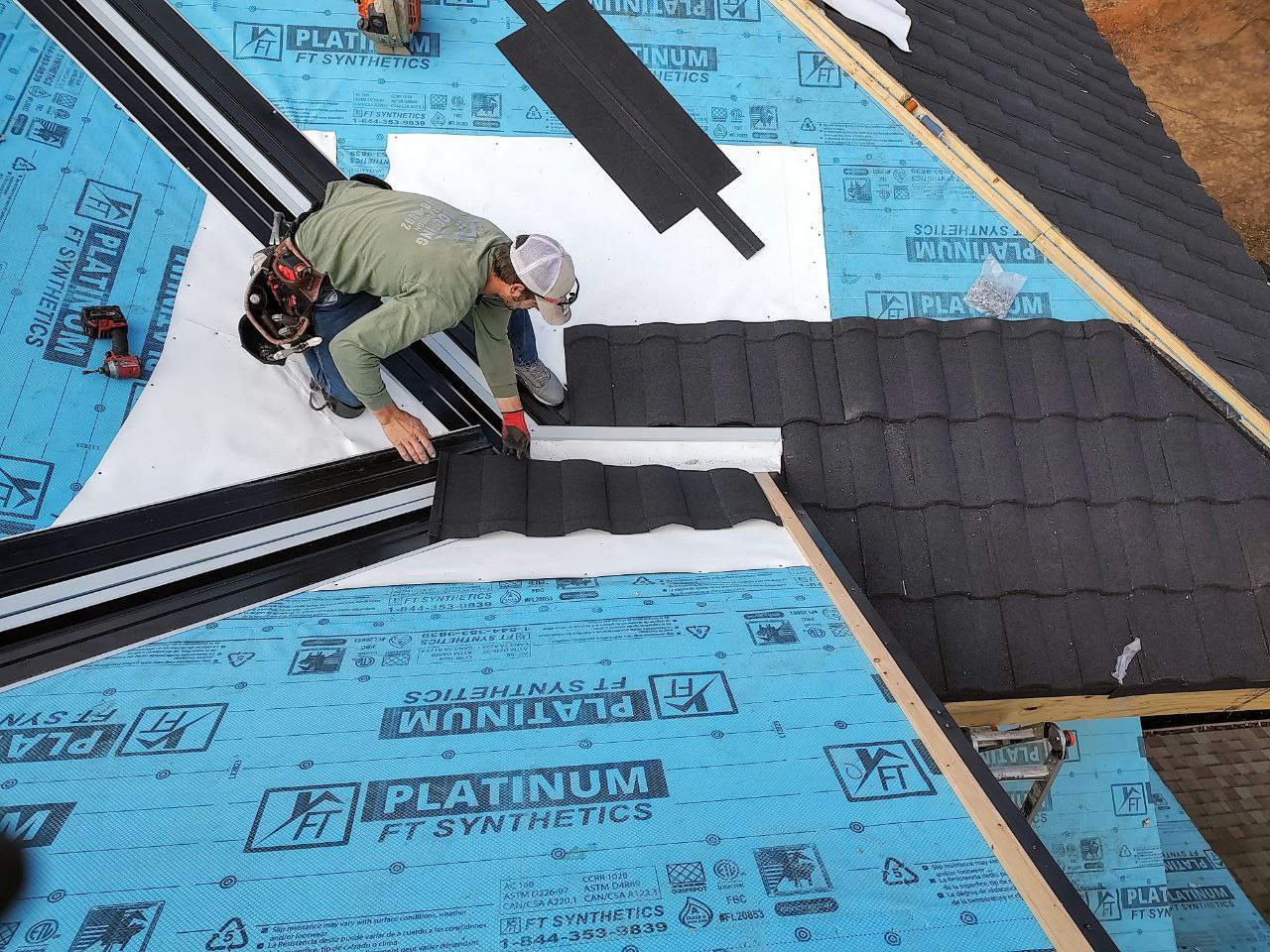 Roofer kneeling on blue synthetic underlayment fastening black metal shingle panels, Dedicated Builders